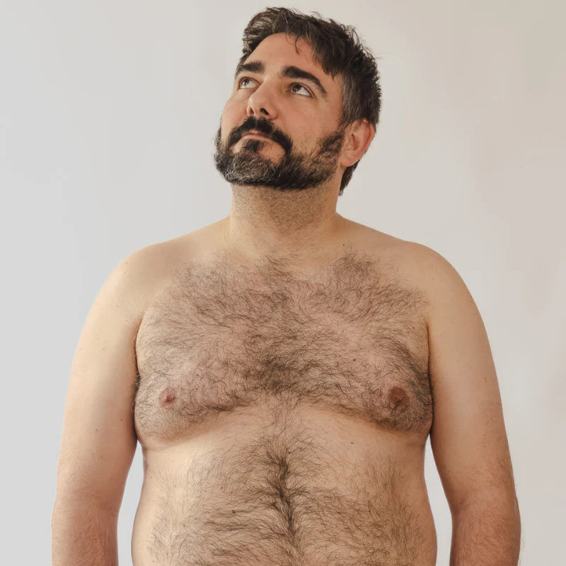 Man with a beard and hairy chest wearing black pants against a plain background