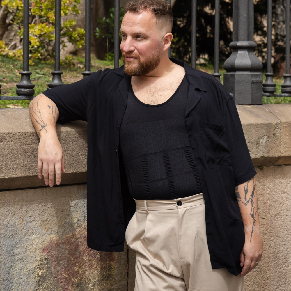 Man wearing a black shirt and beige pants leaning against a stone wall with a metal fence in the background.